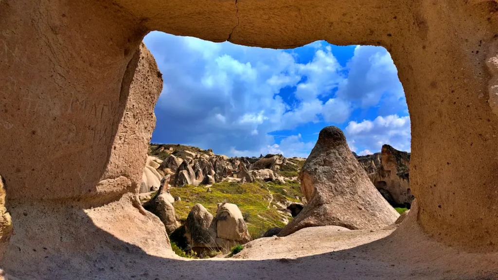 View through a cave opening framing the surreal landscape of Cappadocia, with its dramatic rock formations known as fairy chimneys under a bright blue sky scattered with clouds. The natural arch of the cave creates a window to this unique terrain, dotted with more cave entrances and grassy knolls, capturing the essence of an ancient monastic retreat carved into the earth.
