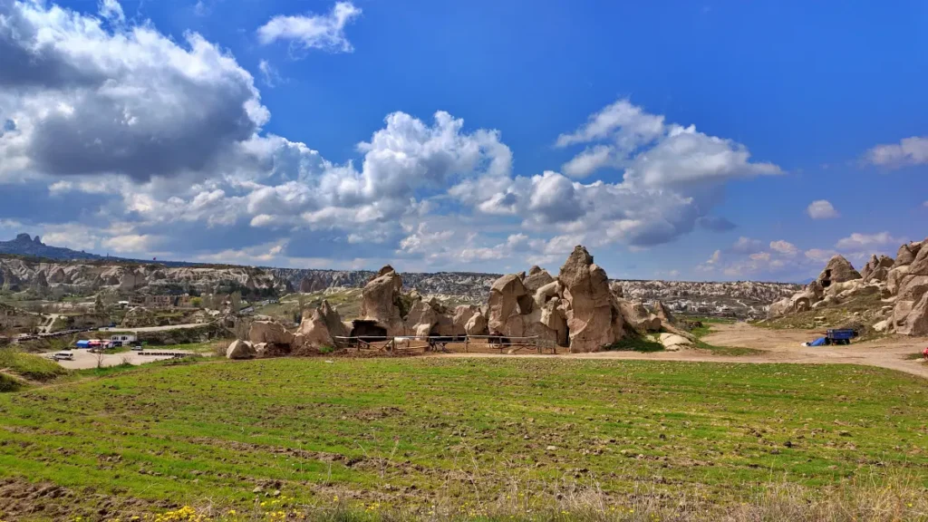 A lush green field in the foreground leads to a cluster of ancient, towering fairy chimneys in the Göreme Open Air Museum under a dynamic sky with billowing clouds. In the middle ground, a few vehicles are parked near the rocky formations, providing a sense of scale to the impressive landscape. The vast expanse of the site extends into the distance, showcasing the unique geological formations that define the terrain of Cappadocia.
