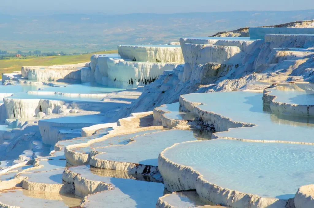 turkey travel A captivating image of Pamukkale's travertine terraces, with their unique, cascading white calcium pools. The photo should showcase this natural wonder's surreal, otherworldly beauty, possibly with the ancient ruins of Hierapolis in the background.