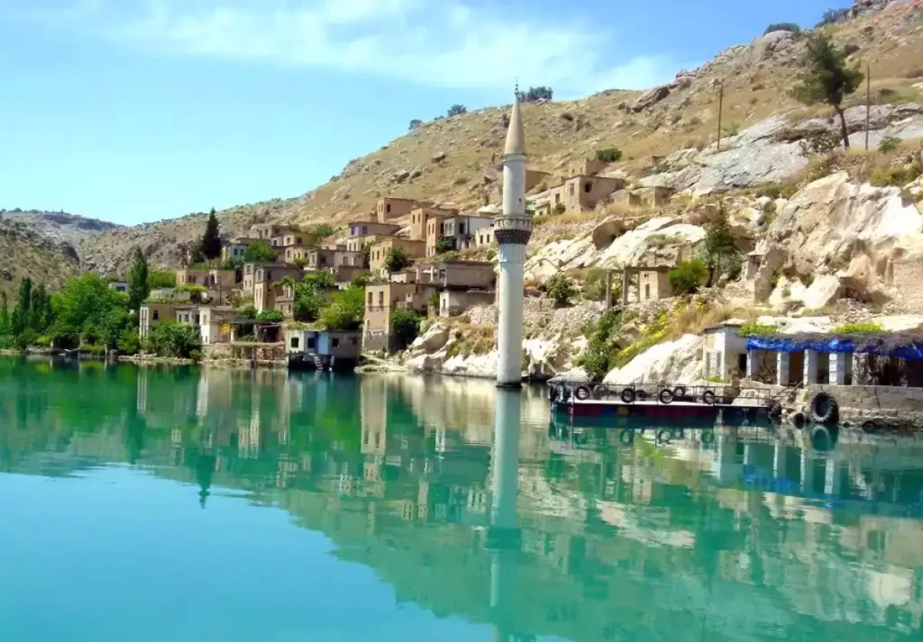 This image depicts the serene and picturesque town of Halfeti, Turkey. The town is known for its partially submerged buildings due to the construction of the Birecik Dam on the Euphrates River. In the photo, traditional stone houses are seen along the water's edge, with their top floors still above water, creating a unique landscape. A prominent minaret stands tall, reflecting the area's rich cultural and historical heritage. The crystal-clear water of the river complements the natural beauty of the surrounding rocky hills, making Halfeti a popular spot for tourists seeking tranquility and scenic beauty.