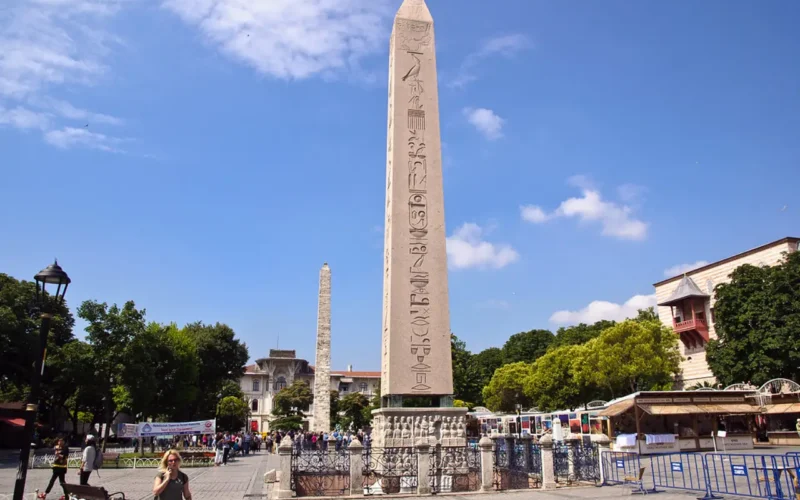 A photo of the Sultanahmet Square in Istanbul, featuring the Obelisk of Thutmose III in the foreground with its hieroglyphic inscriptions clearly visible. Behind it stands the Walled Obelisk, partially obscured by trees. The square is lively with visitors and bordered by low-rise buildings and blue sky with scattered clouds overhead.