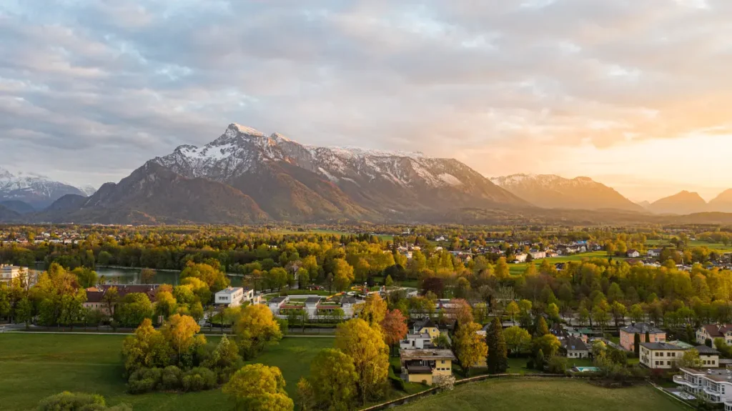 The image presents a breathtaking view of the Untersberg mountain range near Salzburg, Austria, at sunset. The mountain peaks are partially capped with snow and bask in the warm glow of the setting sun. In the foreground, a tranquil expanse of lush green fields and trees in early bloom is interspersed with the roofs of residential buildings, illustrating the harmonious balance between nature and the local community.