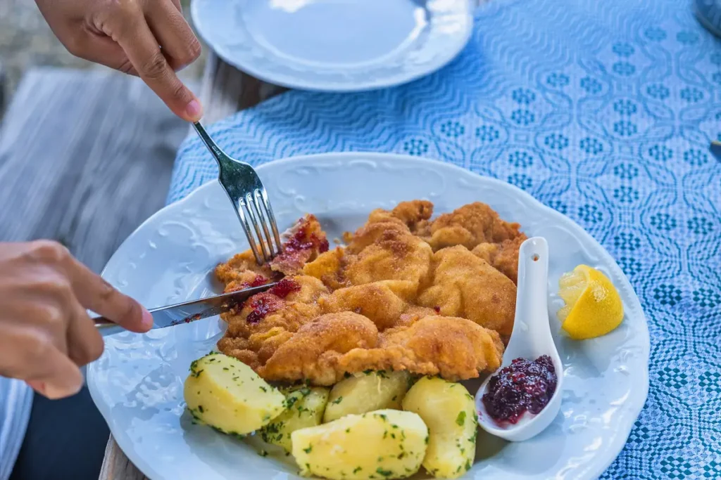 The image shows a plate of traditional Austrian Wiener Schnitzel, accompanied by a side of parsley potatoes and a dollop of lingonberry jam. A person is cutting into the crispy, golden-brown schnitzel with a fork and knife. A wedge of lemon rests in a small white spoon, ready to add a spritz of citrus to the dish. The plate is set upon a blue and white patterned tablecloth, adding a touch of rustic charm to the meal presentation.