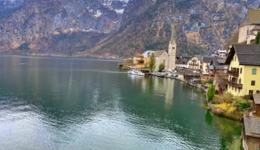 The image depicts a charming lakeside village with European architecture nestled in a mountainous region. The calm body of water reflects the overcast sky and the structures along the shoreline, suggesting a serene, picturesque setting often sought after by travelers and photographers. The town appears to be Hallstatt, a well-known destination in Austria, recognized for its stunning alpine scenery and heritage buildings. This quaint village is a UNESCO World Heritage site and is quite popular among visitors for its traditional houses, historical significance, and natural beauty, including the lake and the Dachstein Alps.