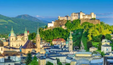 The image displays a panoramic view of Salzburg, Austria, with the Hohensalzburg Fortress prominently situated atop a lush green hill, overlooking the city. Below, the cityscape is dotted with historical buildings, including the Salzburg Cathedral with its distinct green domes and the Franciscan Church's slender spire, set against a backdrop of distant mountains under a clear sky.