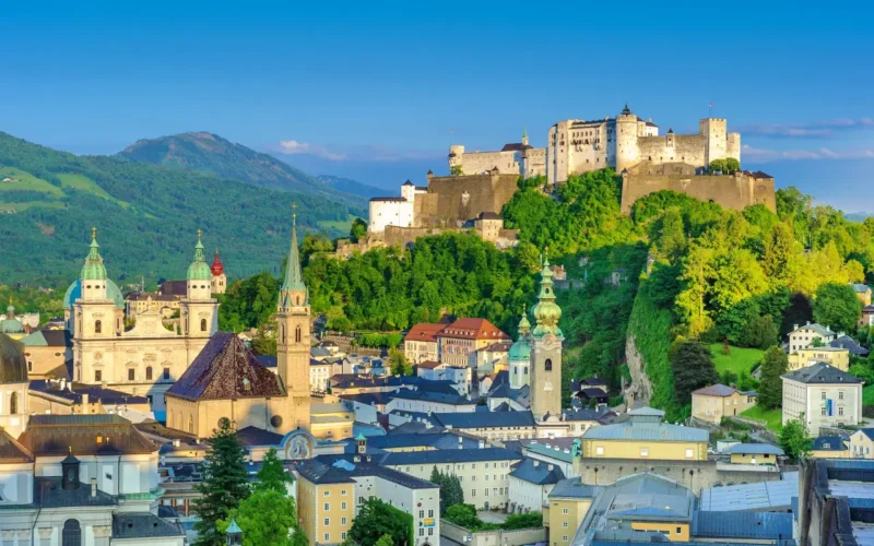 The image displays a panoramic view of Salzburg, Austria, with the Hohensalzburg Fortress prominently situated atop a lush green hill, overlooking the city. Below, the cityscape is dotted with historical buildings, including the Salzburg Cathedral with its distinct green domes and the Franciscan Church's slender spire, set against a backdrop of distant mountains under a clear sky.