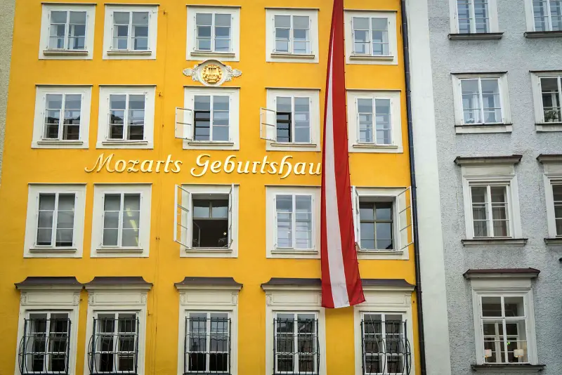 The image shows the vibrant yellow façade of Mozart's birthplace in Salzburg, Austria, with the text "Mozarts Geburtshaus" prominently displayed. White window frames stand out against the bold color, and a large Austrian flag hangs vertically between windows. The building's historical significance is underscored by a crest above the text.