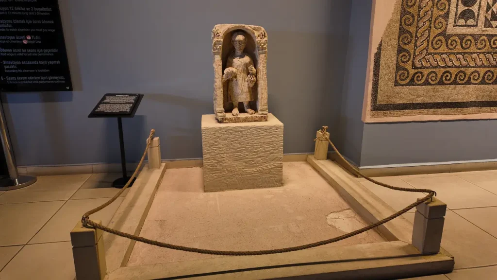 a stone statue displayed at the Zeugma Mosaic Museum. The statue is housed in a small alcove-like frame, depicting a figure holding objects in both hands, possibly fruit or offerings. It is placed on a stone pedestal surrounded by a simple rope barrier to protect the artifact. The background includes an informational plaque, and a mosaic is partially visible on the wall to the right. The setup highlights the care taken to preserve this ancient artifact while offering educational insight to museum visitors.