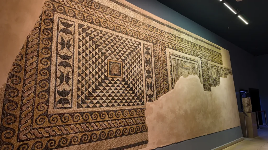 a large, intricately detailed mosaic from the Zeugma Mosaic Museum. The mosaic features geometric patterns with a mix of triangular and swirling designs, framed by decorative borders. The central area showcases a small square panel with a more detailed scene, although parts of the mosaic are missing, leaving sections of the plaster exposed. The craftsmanship demonstrates the artistic precision of Roman-era mosaics, with vibrant earth tones and dynamic shapes. The mosaic is mounted on a wall for display, giving visitors a closer view of this ancient artwork.