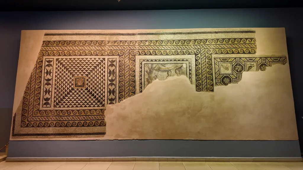 a large, intricately detailed mosaic from the Zeugma Mosaic Museum. The mosaic features geometric patterns with a mix of triangular and swirling designs, framed by decorative borders. The central area showcases a small square panel with a more detailed scene, although parts of the mosaic are missing, leaving sections of the plaster exposed. The craftsmanship demonstrates the artistic precision of Roman-era mosaics, with vibrant earth tones and dynamic shapes. The mosaic is mounted on a wall for display, giving visitors a closer view of this ancient artwork.