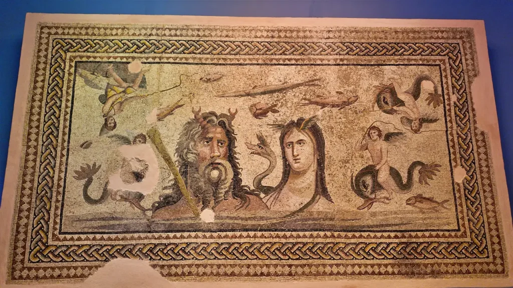 a detailed mosaic from the Zeugma Mosaic Museum, featuring the mythological figures Poseidon, the god of the sea, and Oceanus, the personification of the ocean. Both figures are prominently depicted in the center of the mosaic, surrounded by sea creatures and marine imagery. The surrounding border is intricately designed with interwoven patterns, enhancing the overall beauty of the piece. Some parts of the mosaic have been worn or lost over time, but the craftsmanship and vivid colors still convey a sense of grandeur and mythology