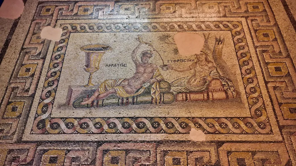 a well-preserved Roman mosaic from the Zeugma Mosaic Museum, depicting two reclining figures, likely mythological or symbolic characters. The inscription next to them reads "AGRATOS" and "EUFRASOYN," indicating possible identities in ancient Greek. One figure holds a drinking vessel, suggesting a banquet or celebration scene. The mosaic's border is richly decorated with geometric and wave-like patterns, emphasizing the craftsmanship involved. Though some portions have been worn away, the vivid colors and detailed imagery reflect the grandeur of Roman art and social life