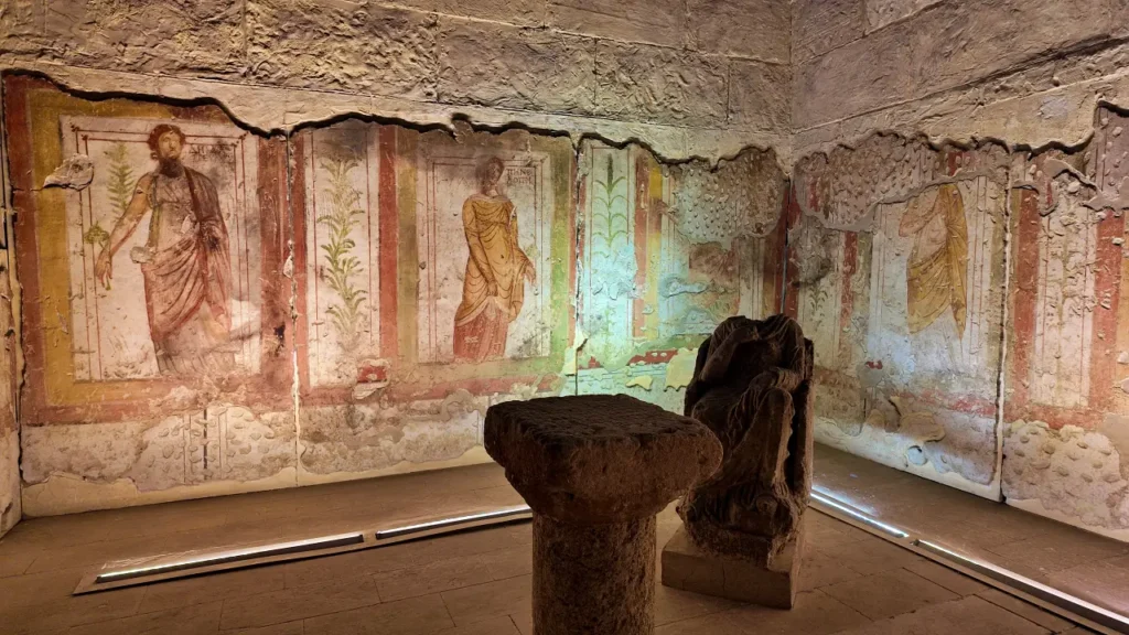 a room from the Zeugma Mosaic Museum, featuring beautifully preserved wall frescoes. These frescoes depict figures draped in classical garments, likely from Roman times, surrounded by architectural and nature motifs. The walls have been worn with time, but the vivid colors and intricate designs are still visible. In the center of the room, two stone artifacts are displayed, possibly parts of columns or altars, adding to the room's historical significance. The lighting emphasizes the textures and colors of both the frescoes and the stone relics, transporting viewers back to the grandeur of ancient Roman interiors