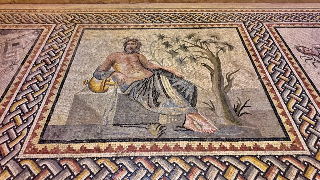 a detailed mosaic from the Zeugma Mosaic Museum, depicting a reclining male figure, possibly a river god or mythological figure. He is shown lounging on a stone structure, holding a golden vessel, with a tree in the background. The mosaic is framed by a complex geometric border, showcasing expert craftsmanship with interwoven patterns. The vivid colors and intricate details, such as the folds of the figure’s robe and the naturalistic depiction of his surroundings, highlight the artistic achievements of the Roman-era mosaics