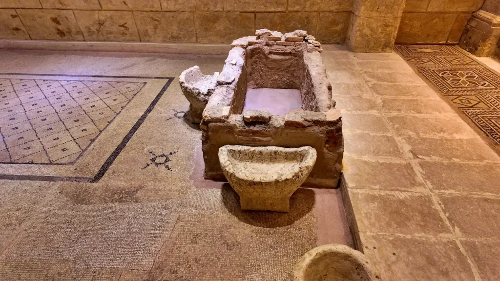 an ancient stone sarcophagus or basin displayed at the Zeugma Mosaic Museum. The rectangular structure is weathered, with visible signs of age and wear. The basin or sarcophagus is accompanied by smaller stone artifacts placed around it. It rests on a mosaic floor featuring geometric patterns, further emphasizing the historical context of the display. The surrounding space showcases more Roman-era mosaic designs, blending the architectural and artistic achievements of the ancient city of Zeugma. This artifact likely played a ceremonial or functional role in ancient times