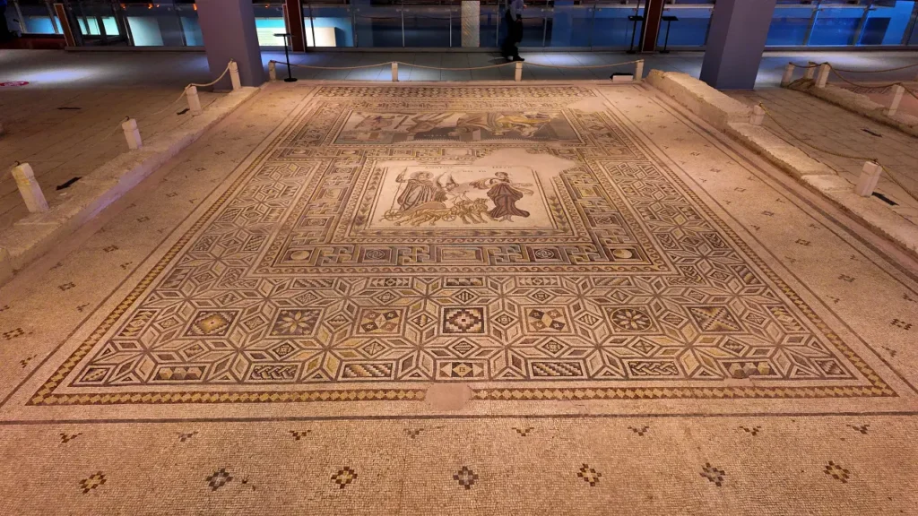 mosaic floor from the Zeugma Mosaic Museum, showcasing intricate geometric patterns framed by decorative borders. The center of the mosaic contains a detailed scene featuring two human figures, likely mythological or historical, accompanied by a chariot and horses. The surrounding geometric designs are meticulously crafted, with star-like patterns and colorful tile arrangements adding depth to the piece. The overall mosaic is framed by stone walls, indicating its preservation as part of an ancient Roman villa or public building. The exhibit is well-lit, allowing visitors to fully appreciate the artistry and scale of this ancient masterpiece